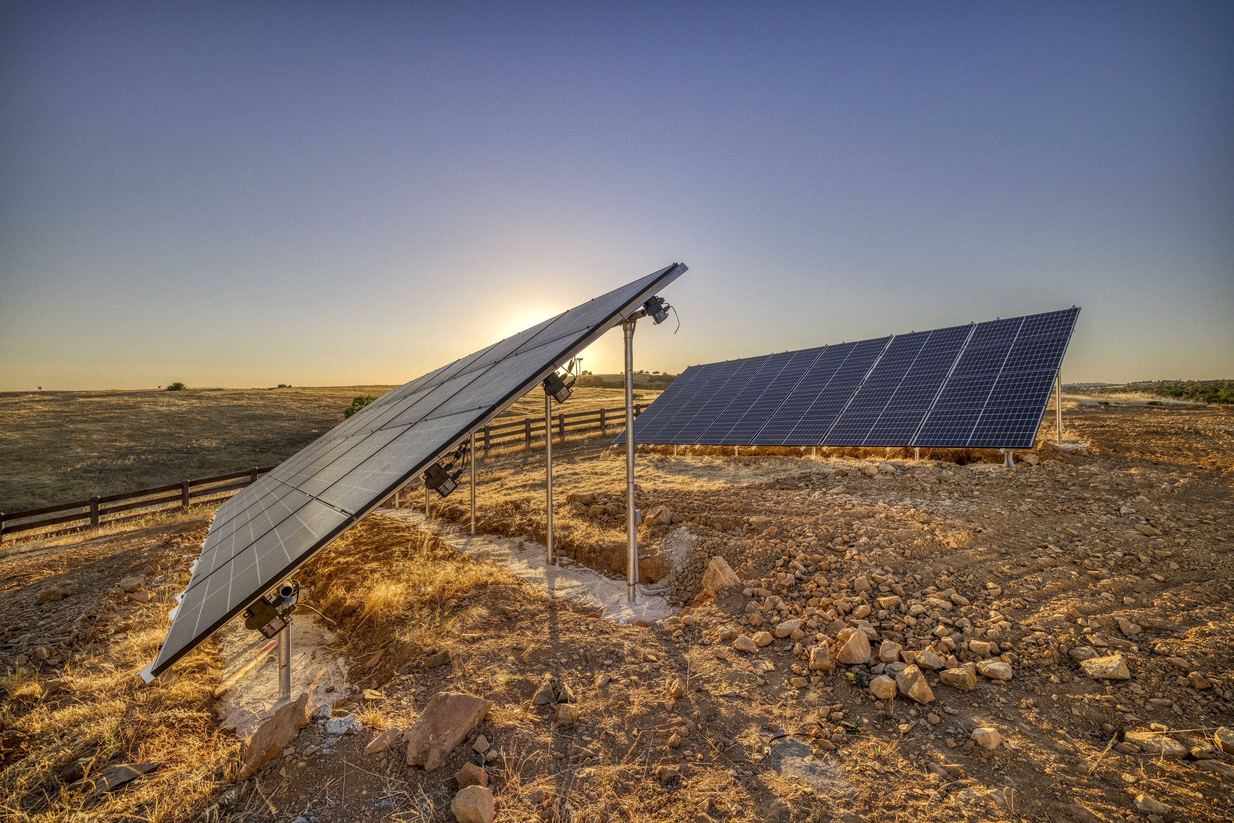 Solar panels installed in a rural area at sunset, with a wooden fence in the background and a clear sky.