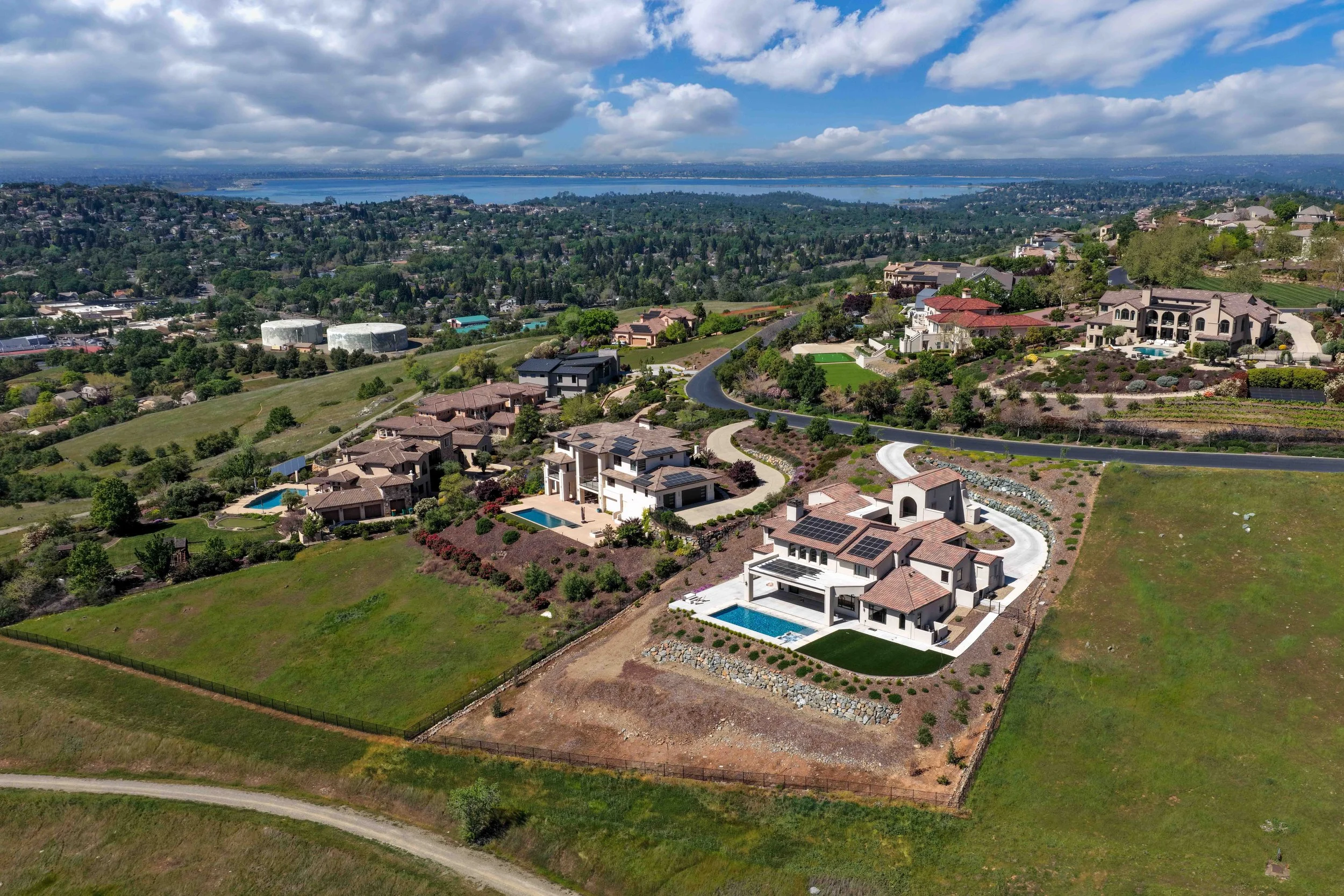 An aerial view of a suburban neighborhood with large houses, some with pools, situated on hilly terrain with winding roads. In the background, there is a body of water and a distant city skyline under a partly cloudy sky.