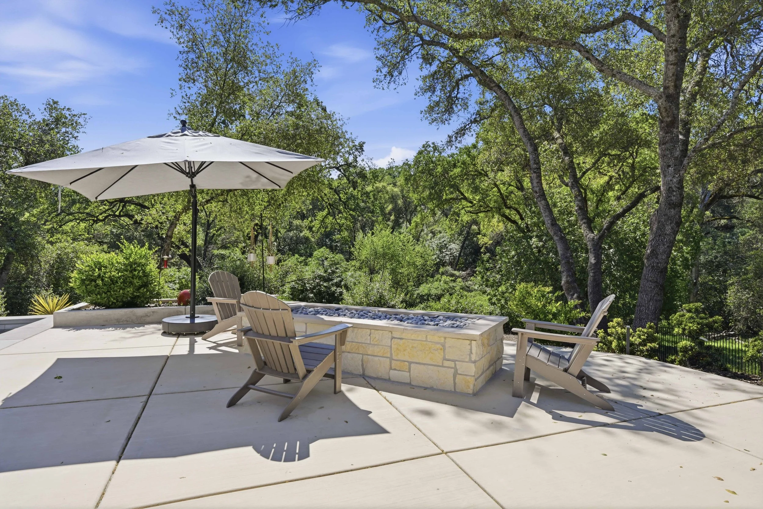 A backyard patio with wooden chairs and a white umbrella, surrounded by trees and greenery with a clear blue sky.