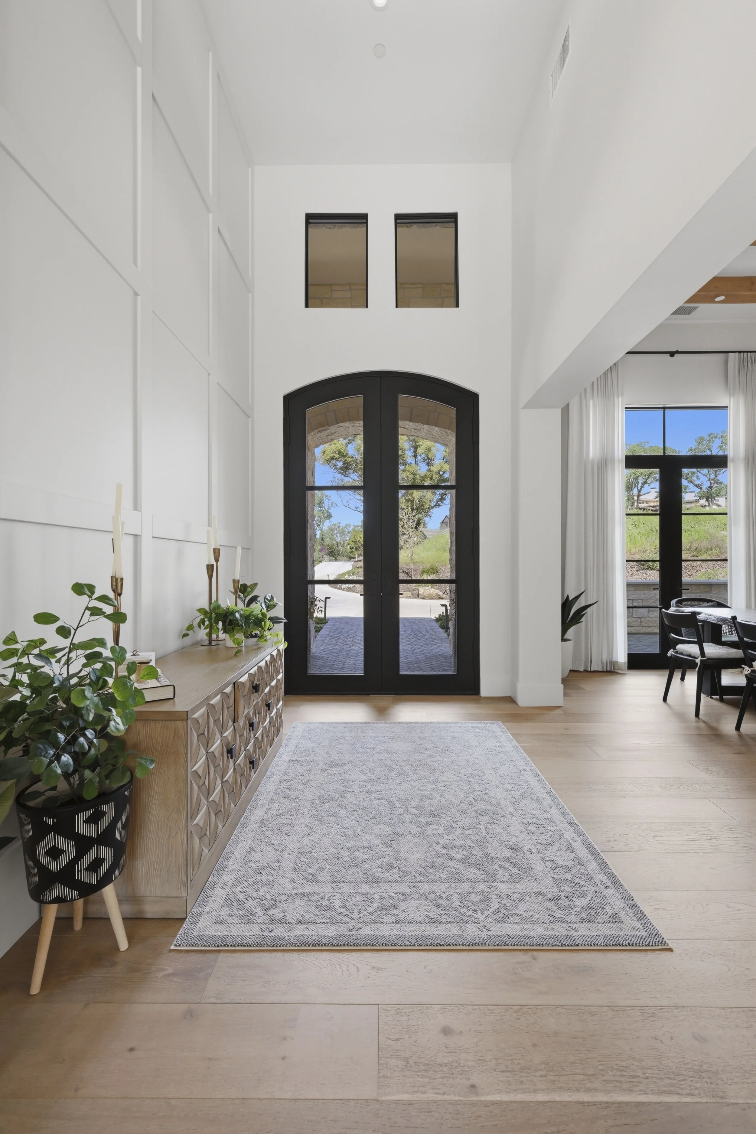 Modern entryway with black arched front door, white walls, light wood flooring, and large windows with white curtains. Potted plants and a decorative sideboard are on the left, and a gray area rug covers part of the floor.