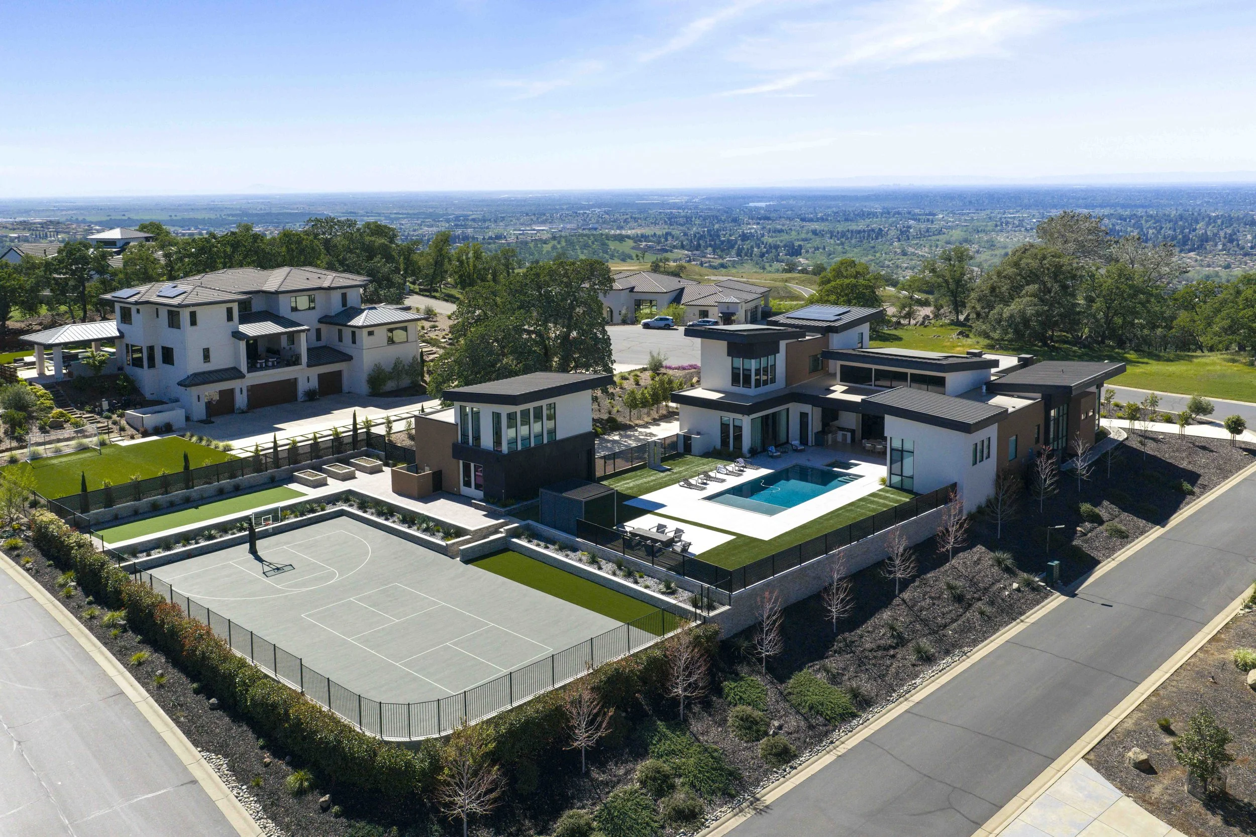 Aerial view of modern residential area with large houses, a swimming pool, tennis court, and green landscaping, overlooking a valley and distant cityscape under a clear blue sky.