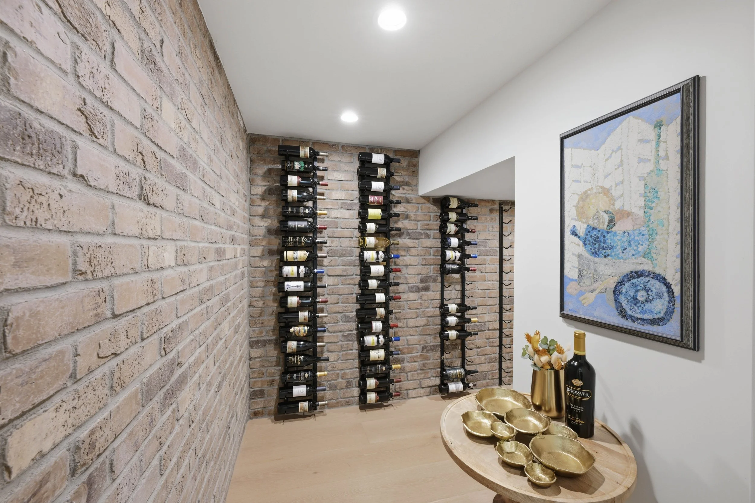 A wine cellar with a brick wall, multiple wine bottles stored on wall-mounted racks, a modern abstract painting on the white wall, and a wooden table with gold decorative bowls and champagne bottles.