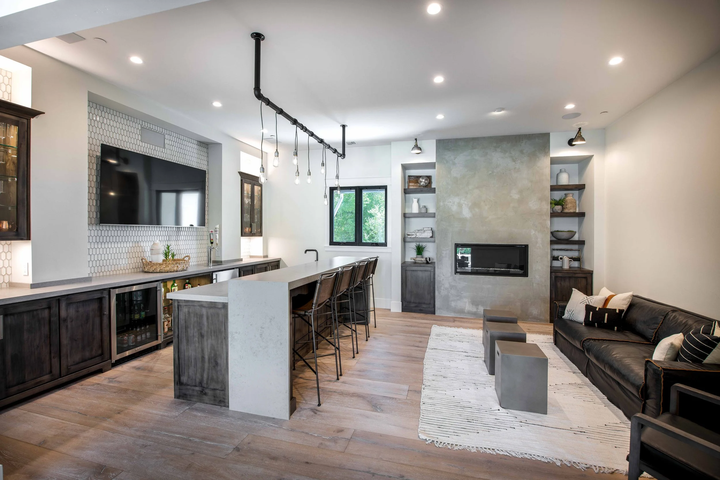 Modern living room and kitchen area with hardwood floors, white walls, and ceiling. The kitchen has a dark wooden cabinet with a glass door and a white countertop island with bar stools. A flat-screen TV is mounted on a wall with white honeycomb tile