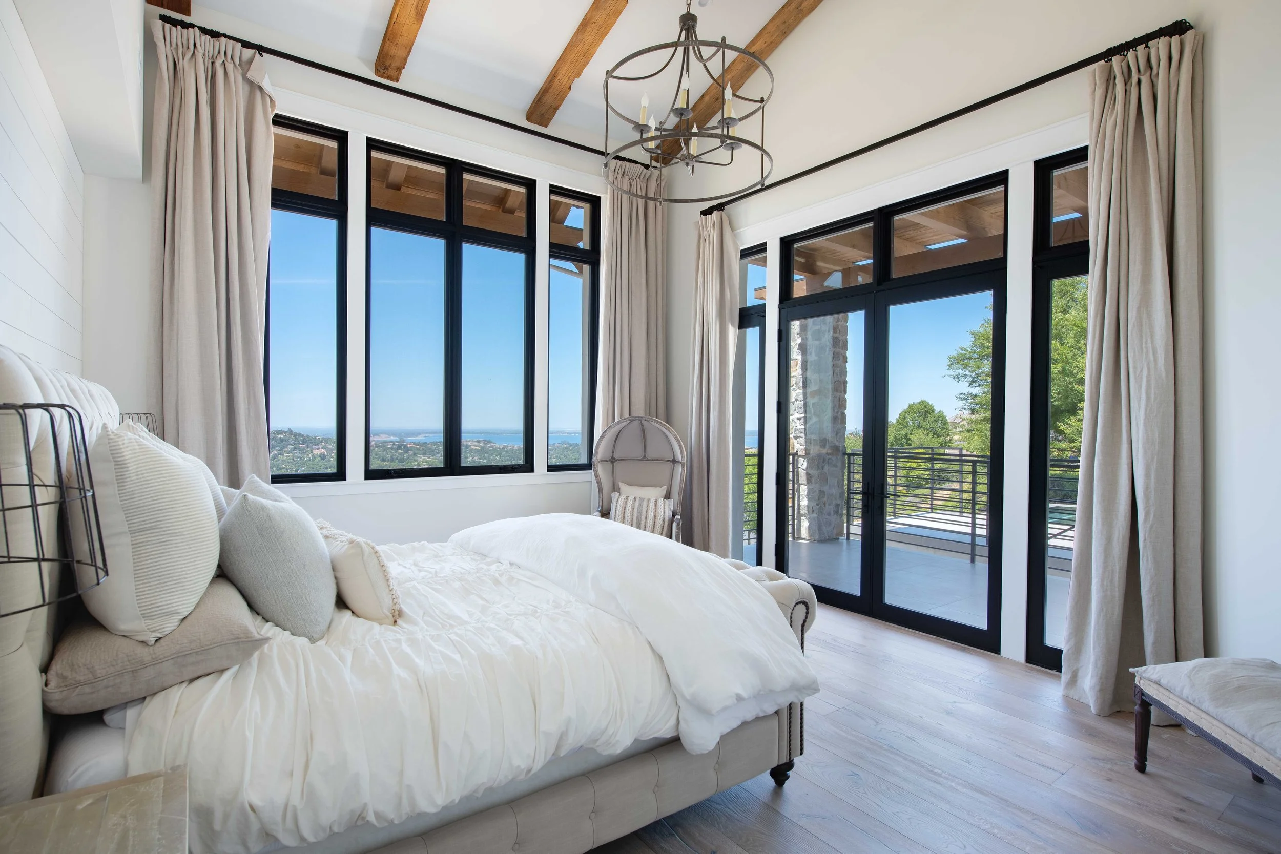 Bedroom with large windows showing a view of trees and water, cream-colored curtains, a chandelier, a white bed with pillows, a small light-colored chair, and a wooden bench.
