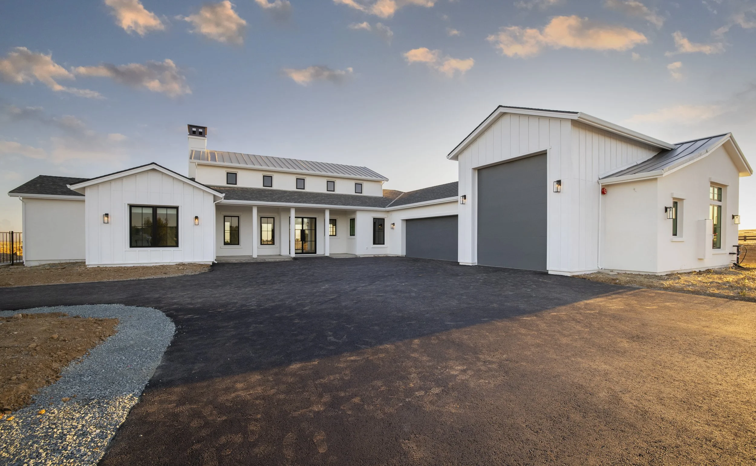 Newly constructed white house with gray garage doors, black window frames, and a dark asphalt driveway, under a partly cloudy sky during sunset.