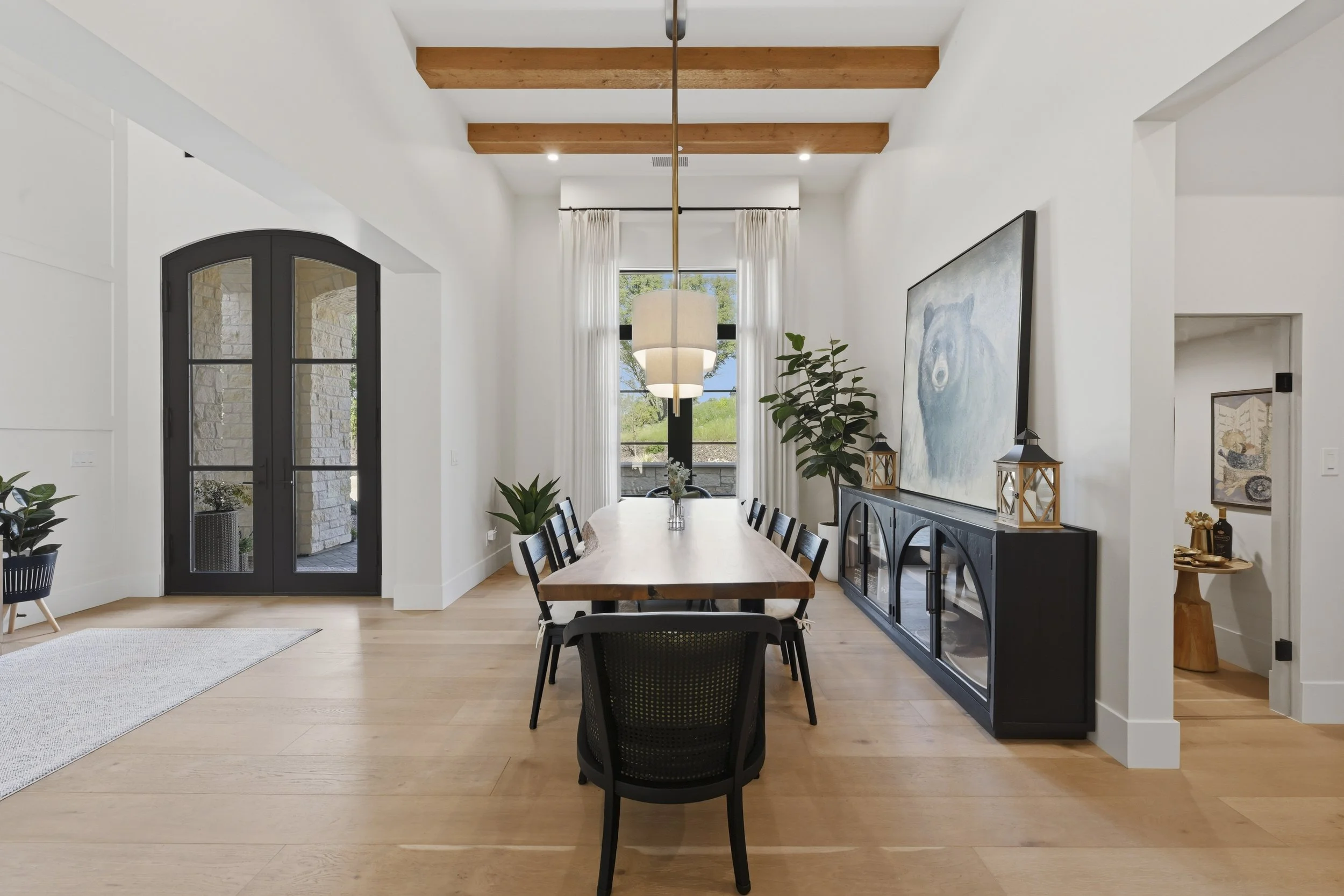 A modern dining room with a wooden table and black chairs, large window with white curtains, artwork on the wall, potted plants, and decorative lanterns on a sideboard, with wooden ceiling beams and double glass doors leading outside.
