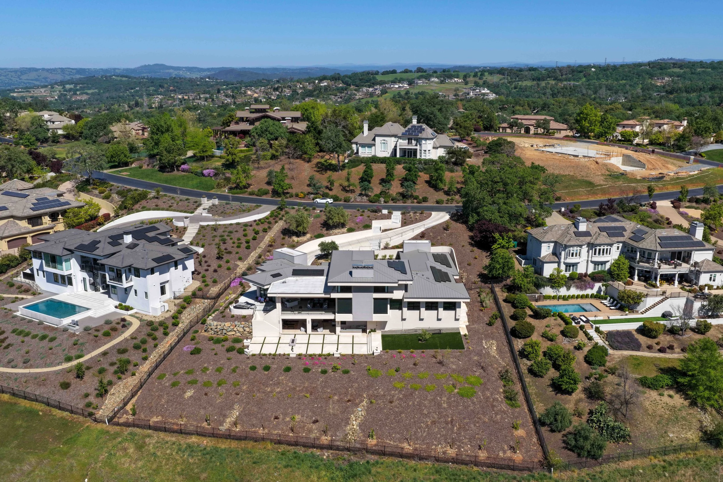 Aerial view of a suburban neighborhood with modern houses, some with solar panels, swimming pools, landscaped yards, and a winding road, surrounded by greenery and rolling hills in the background.