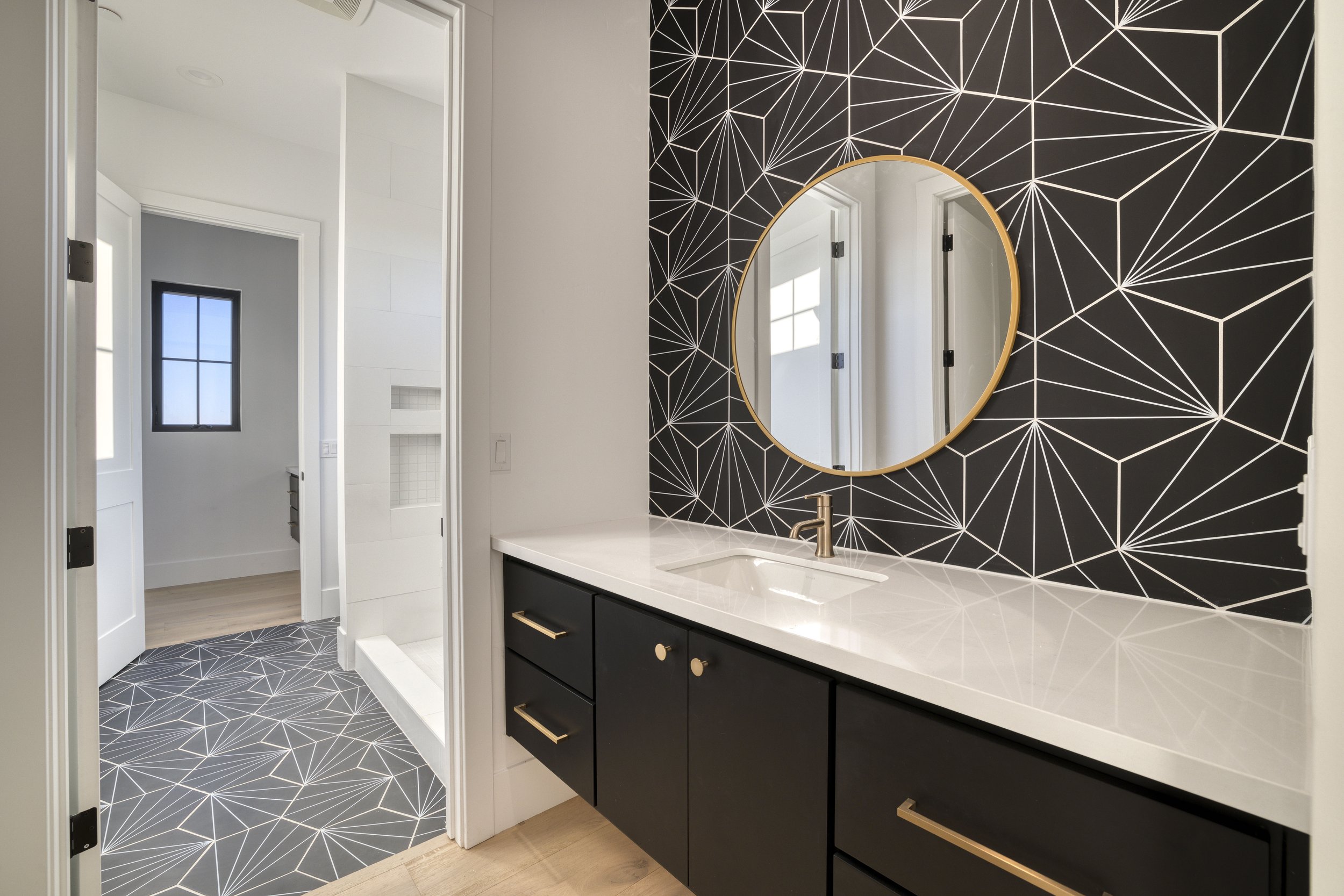 Modern bathroom with black geometric patterned wallpaper behind a large oval mirror, black vanity with gold handles, white countertop, and gold faucet. Light wood flooring transitions to black geometric patterned tiles in the shower area visible thro