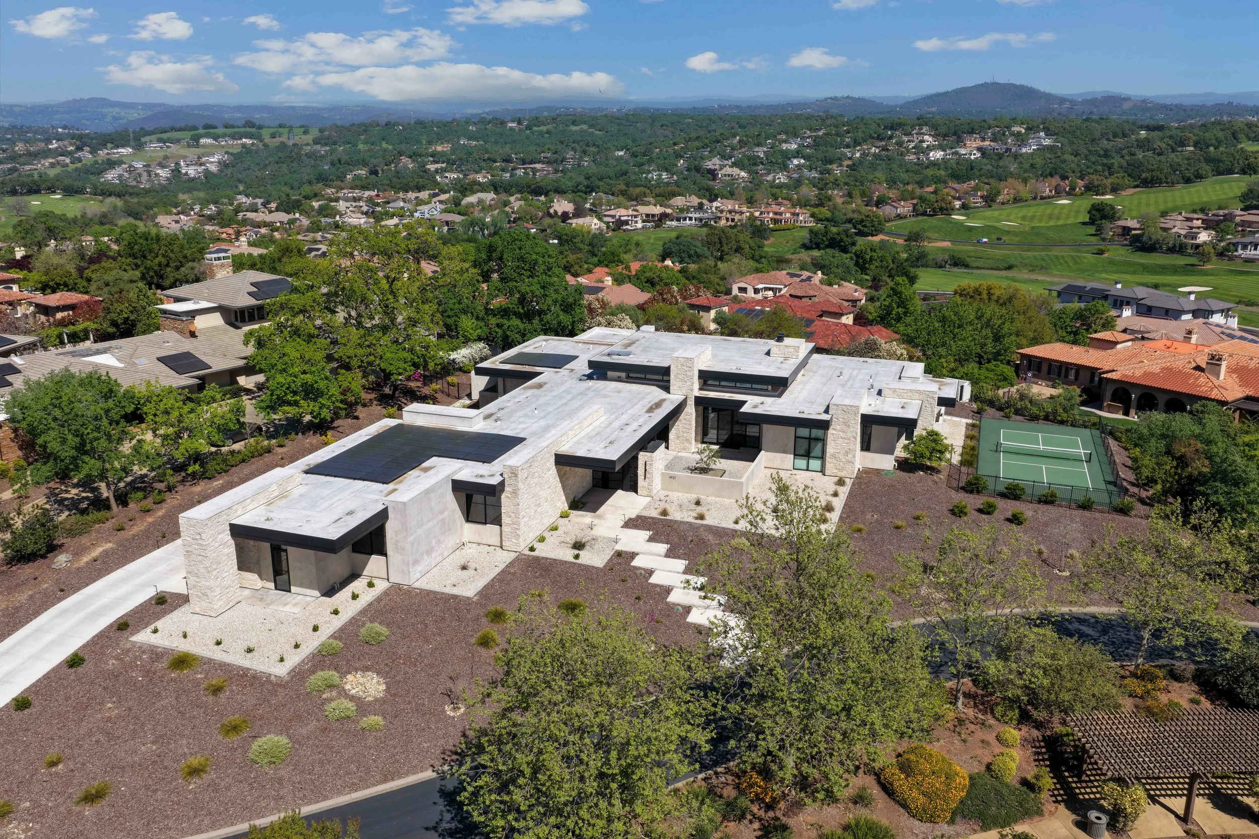 Aerial view of a suburban neighborhood with modern houses, green trees, a golf course, and rolling hills in the background under a partly cloudy sky.