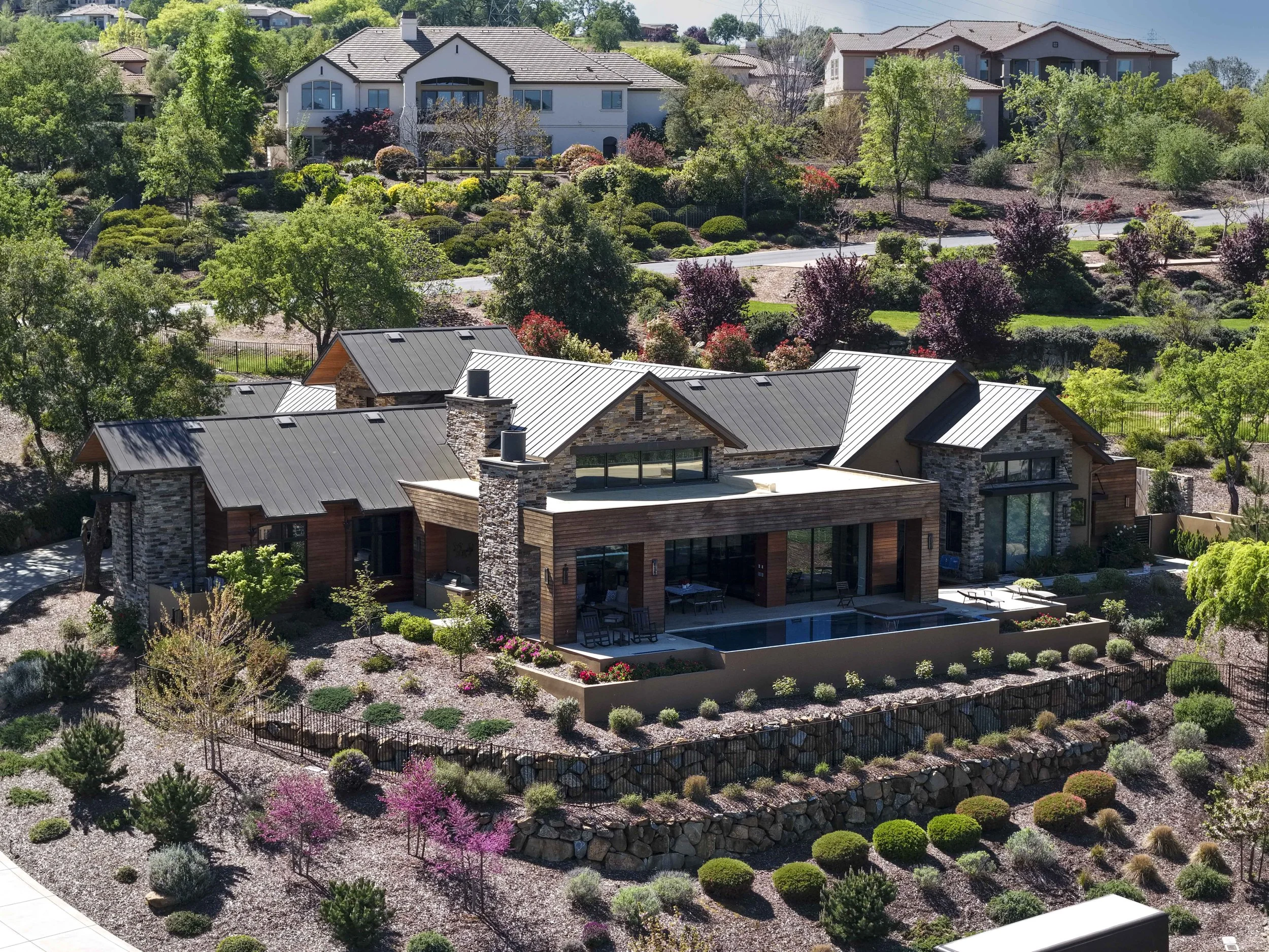 Aerial view of a residential neighborhood with modern houses, lush green trees, and well-maintained landscaping including colorful flowering trees and bushes.