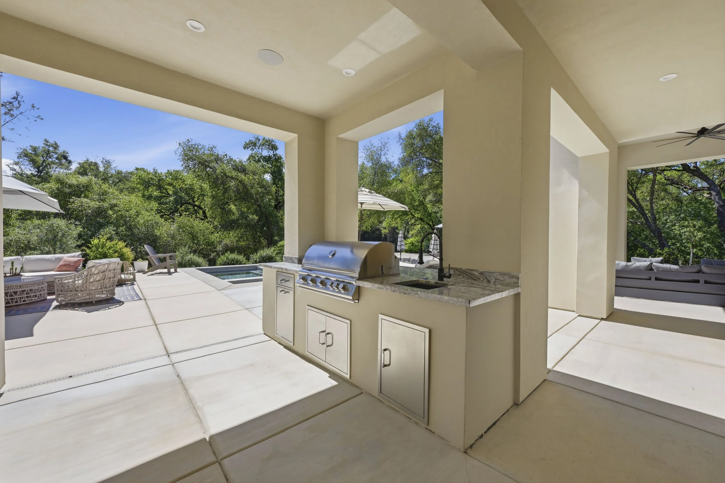 Outdoor patio area with a built-in gas grill, seating, umbrellas, and surrounding trees on a sunny day.