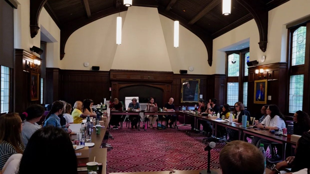 A large conference room with high dark wood ceilings and walls, filled with people seated around long rectangular tables, participating in a meeting or discussion.
