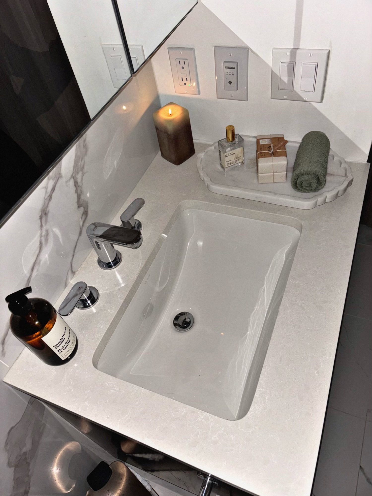 Bathroom countertop with a white sink, silver faucet, brown soap dispenser, candle, perfume bottle, wrapped soap, rolled towel, and electrical outlets with various switches and outlets.