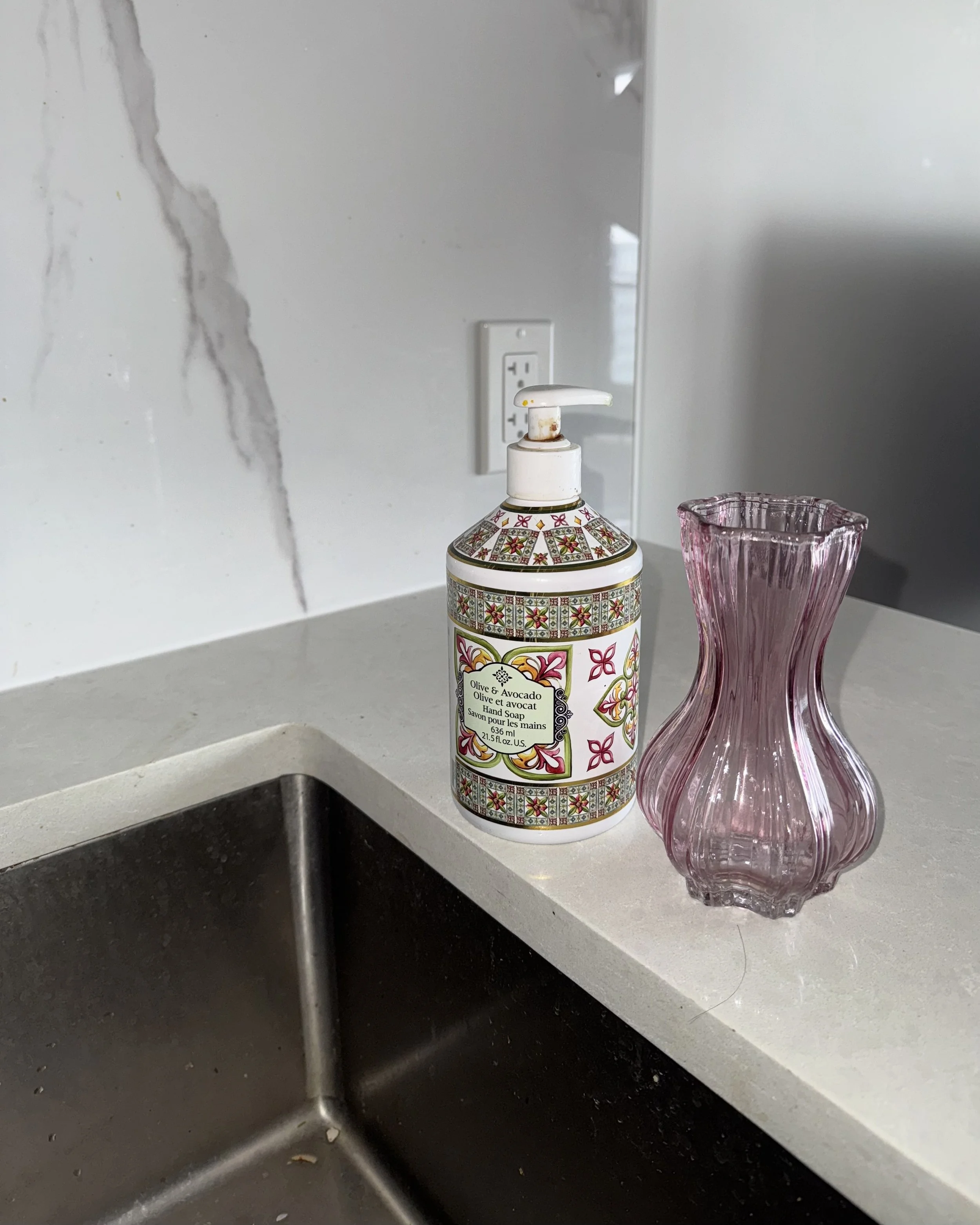 A decorative bottle of olive and avocado hand soap next to a pink glass vase on a kitchen countertop, with a marble backsplash and electrical outlet in the background.