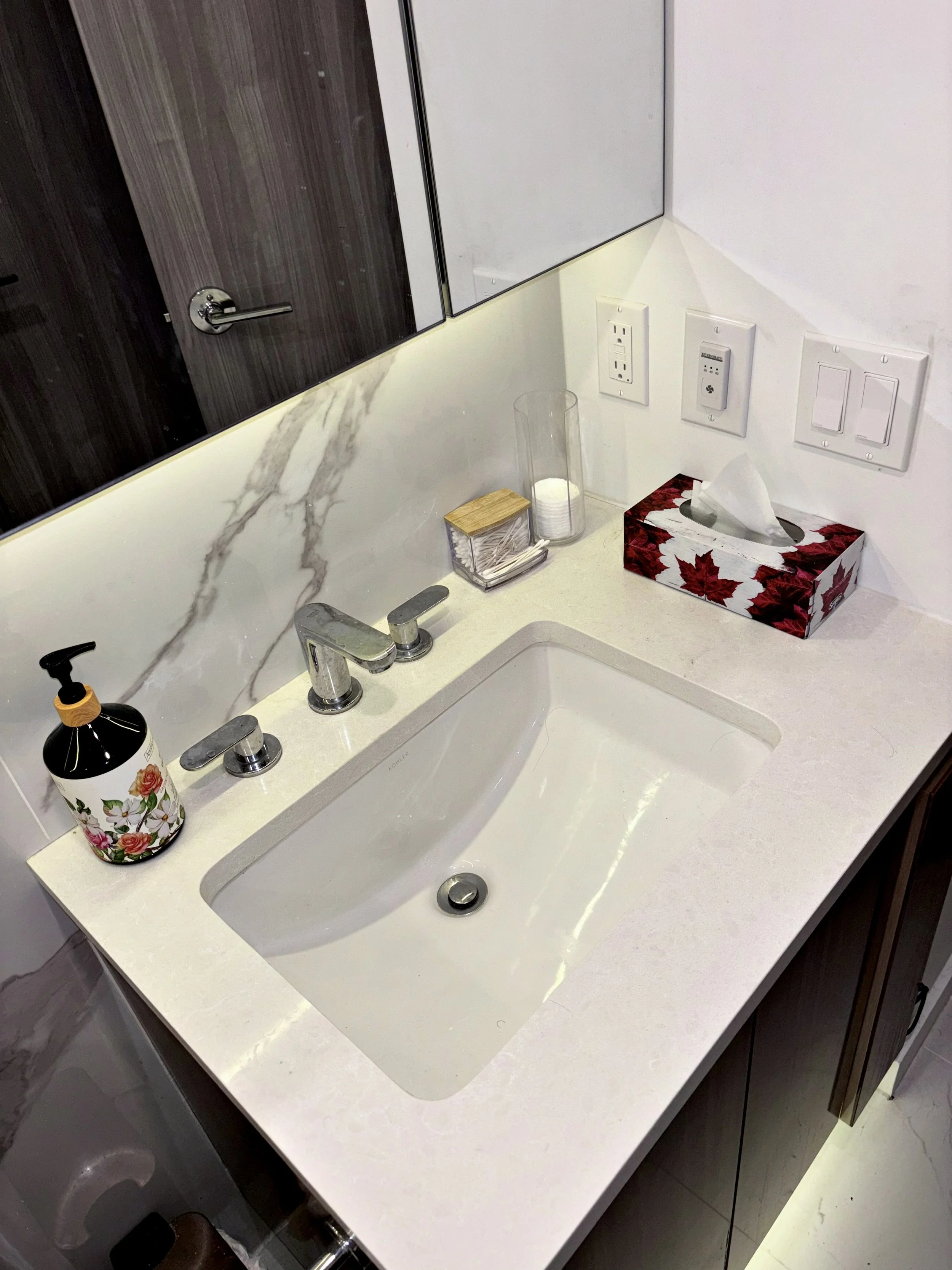 Bathroom sink with faucet, soap dispenser, tissue box, and various electrical outlets and switches in the background.
