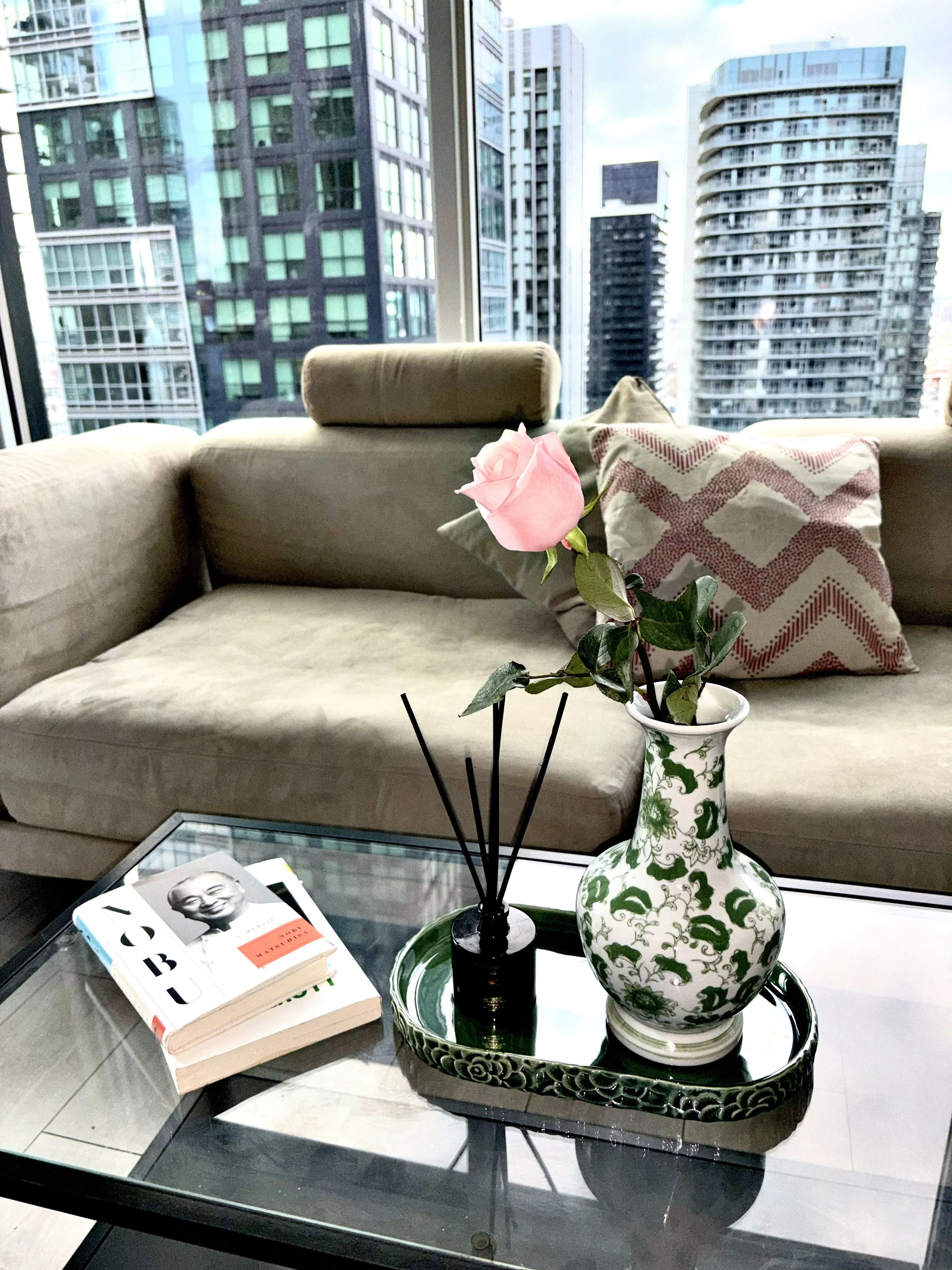 Living room with a beige sofa, a pink rose in a decorative green and white vase, a black reed diffuser, a book on the glass coffee table, and a cityscape view of tall buildings through large windows.
