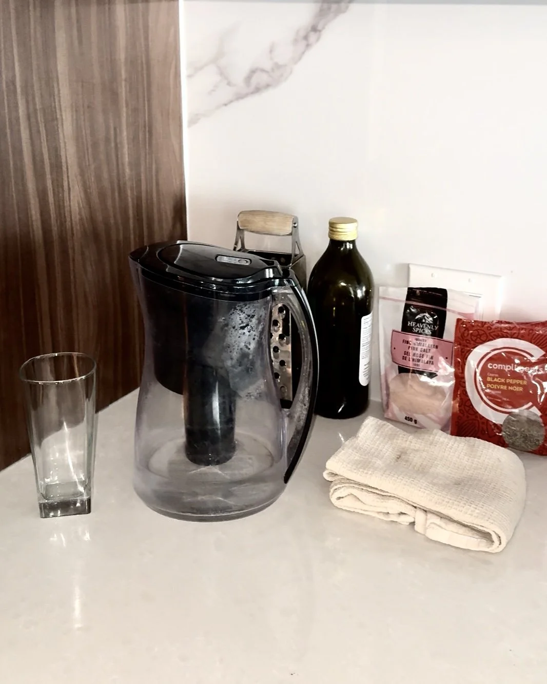 A kitchen countertop with a glass, a glass water filter pitcher, a bottle of black salt, a bag of pink Himalayan salt, a bag of black pepper, a beige dish towel, and part of a wooden chair in the background.