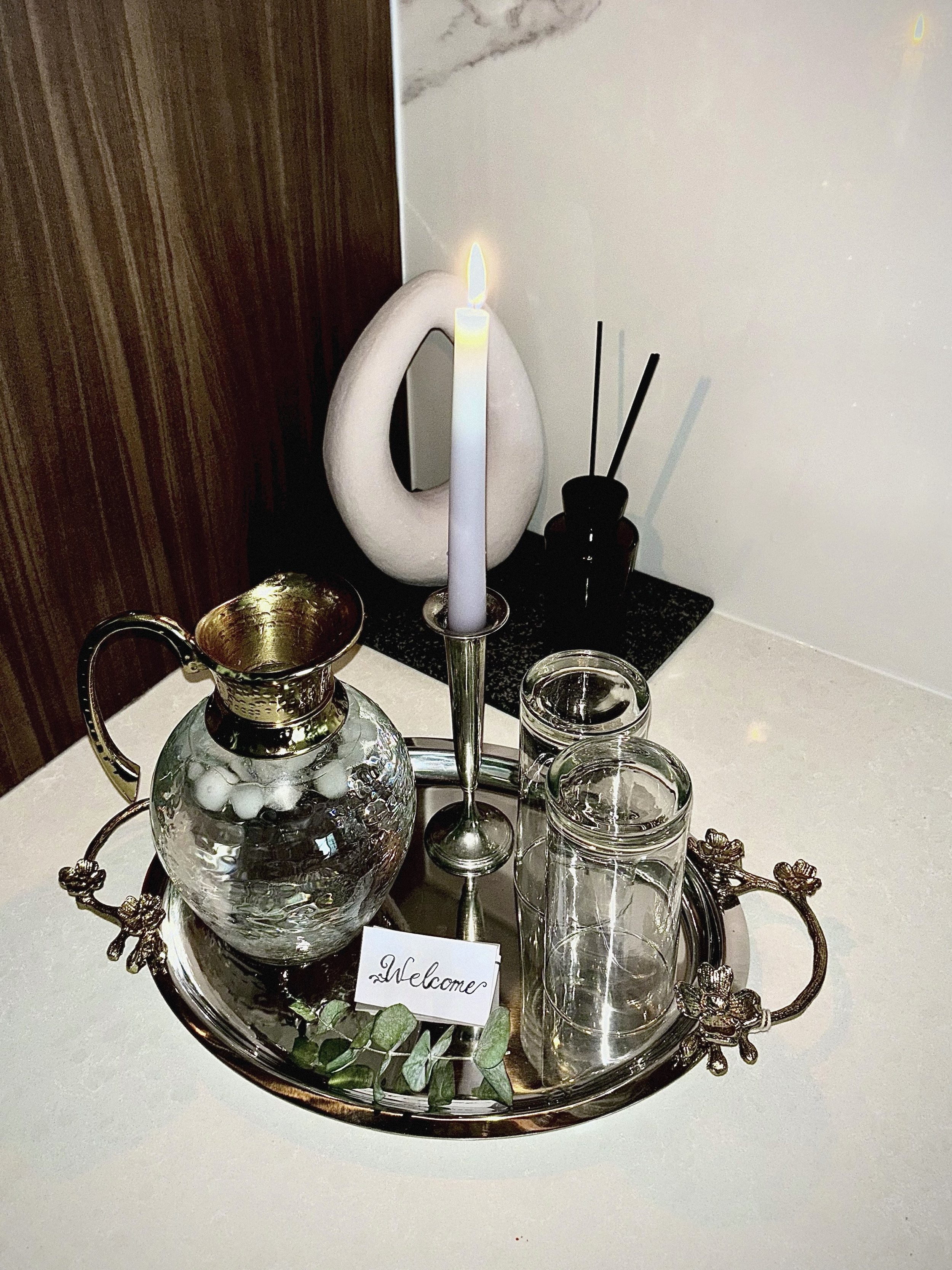 A decorative table setup featuring a silver tray with a glass fish-shaped pitcher, two empty glasses, and a paper note that says "Welcome," with a silver candle holder and a lit candle, and a white artistic sculpture in the background.