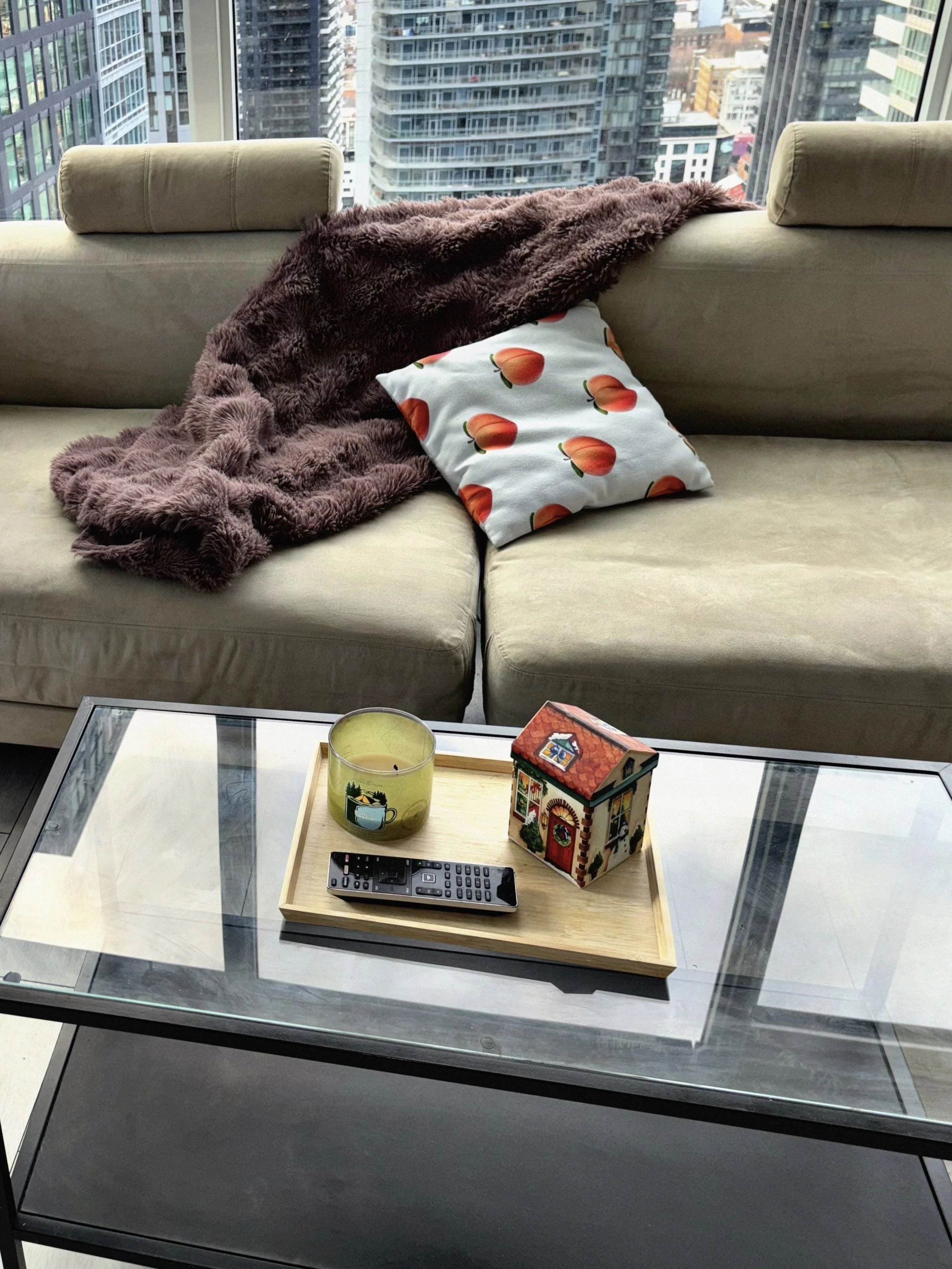 A living room with a beige sofa, a brown fuzzy blanket, and a pillow with a peach pattern. In front, a glass coffee table with a wooden tray holding a candle, a remote control, and a decorative house.