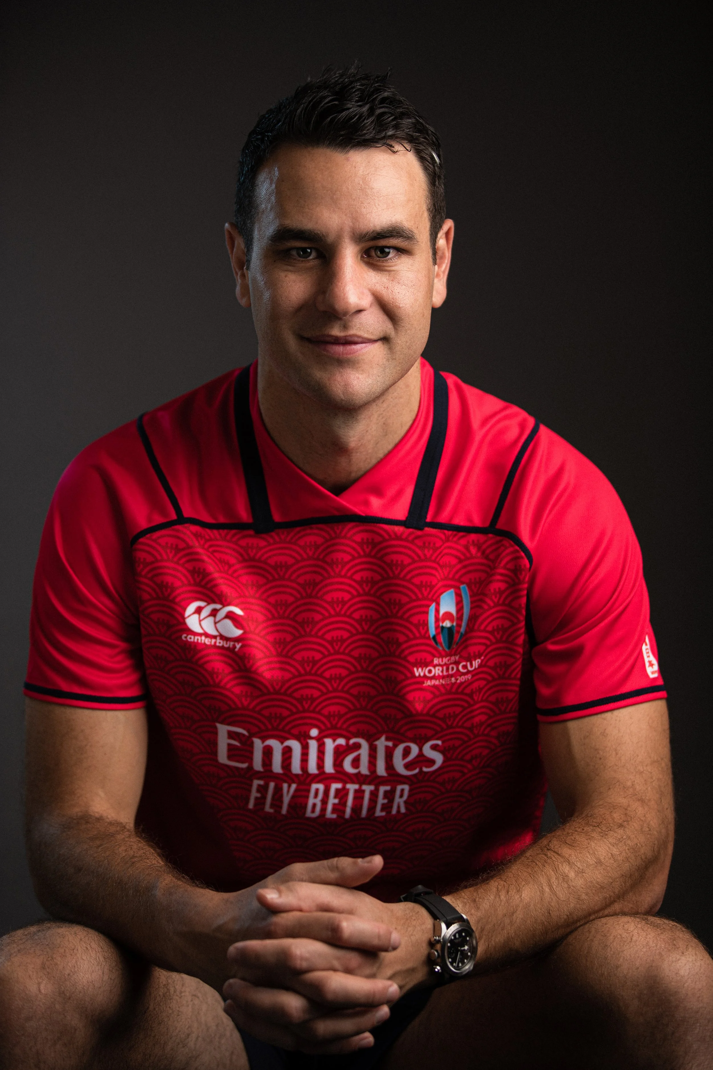 Ben O'Keeffe in a red rugby jersey with black accents, sitting against a dark background, smiling slightly, with his hands clasped.