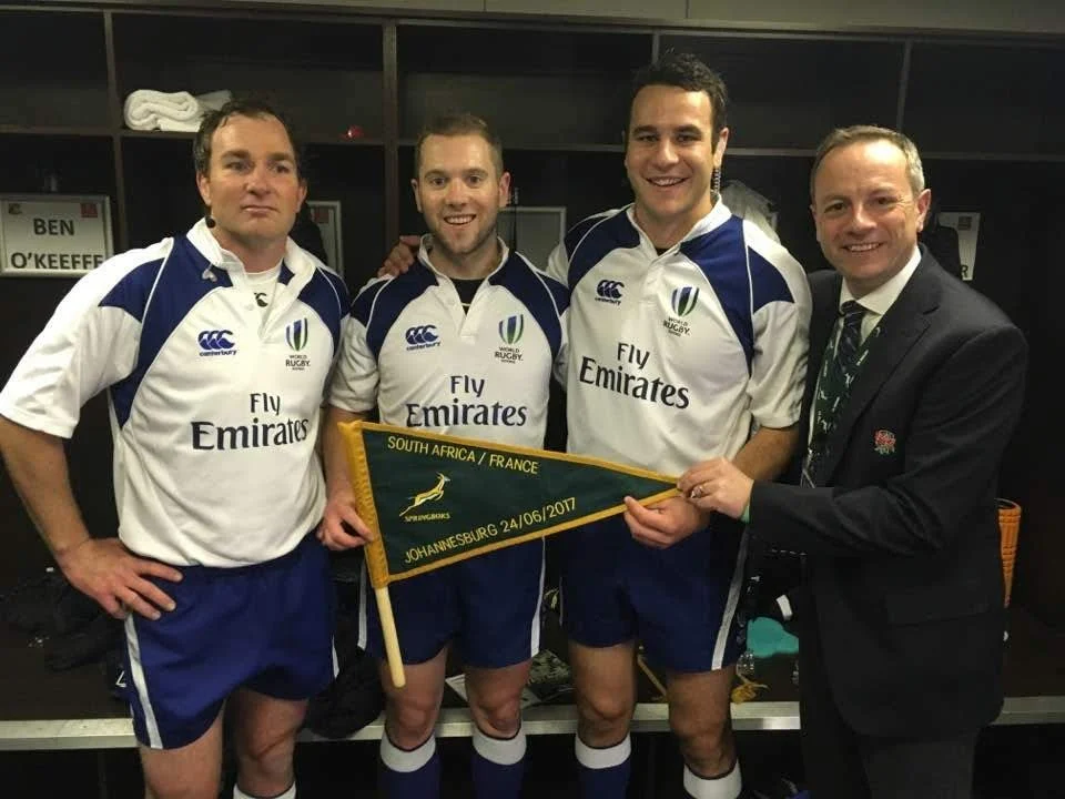Four rugby referees, including Ben O'Keeffe, in team jerseys standing together with a man in a suit holding a banner that reads 'South Africa / France, Johannesburg, 24/06/2017' in a locker room.