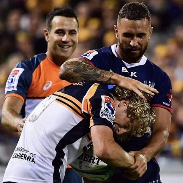 Three rugby players involved in a tackle during a match, with two players in blue jerseys and one in a white and orange jersey. The stands with spectators are blurred in the background and includes referee Ben O'Keeffe .