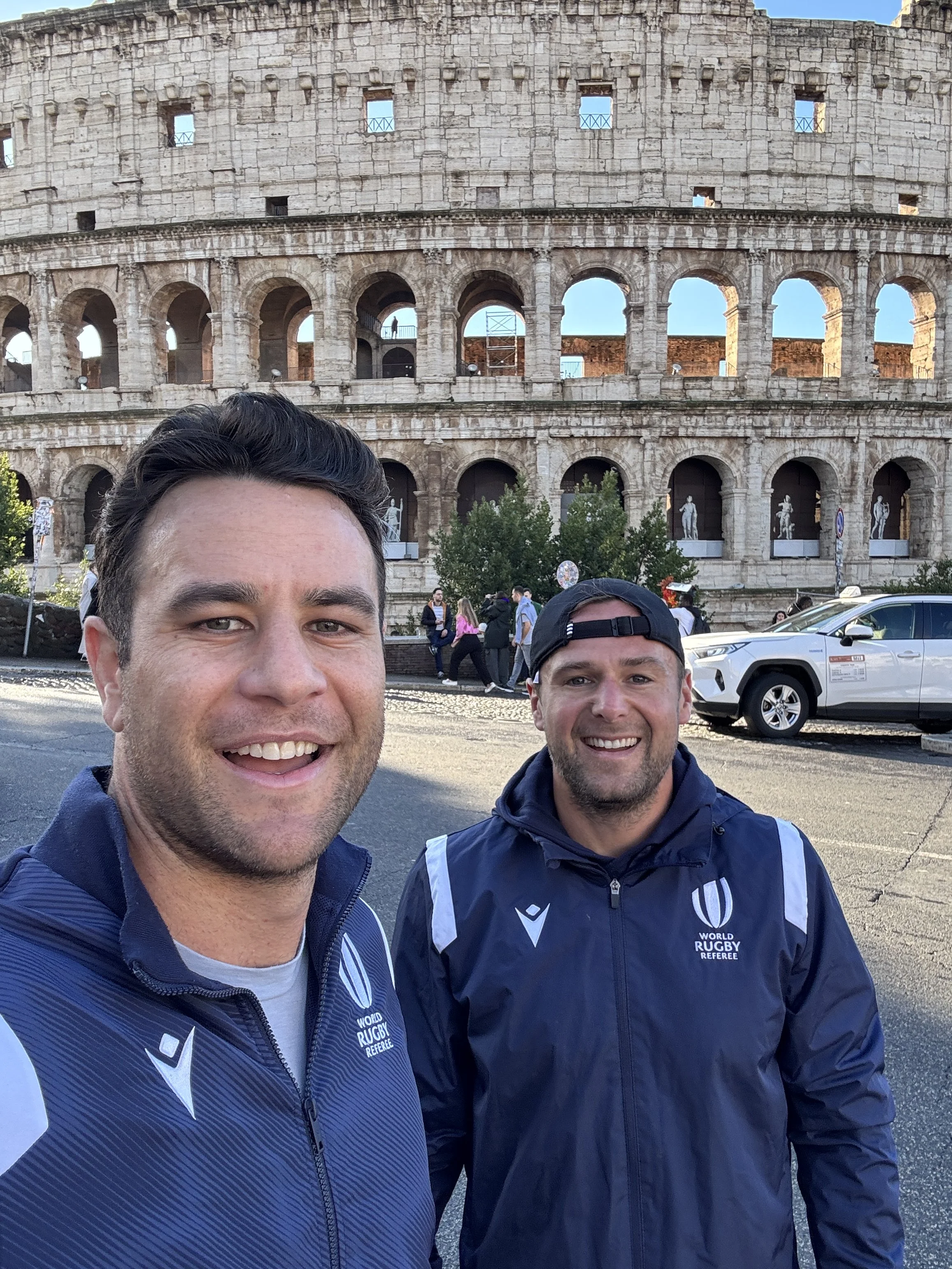 Two men smiling and taking a selfie in front of the Colosseum in Rome, Italy; one wearing a World Rugby referee jacket and the other wearing a black cap are referees Ben O'Keeffe and James Doleman.