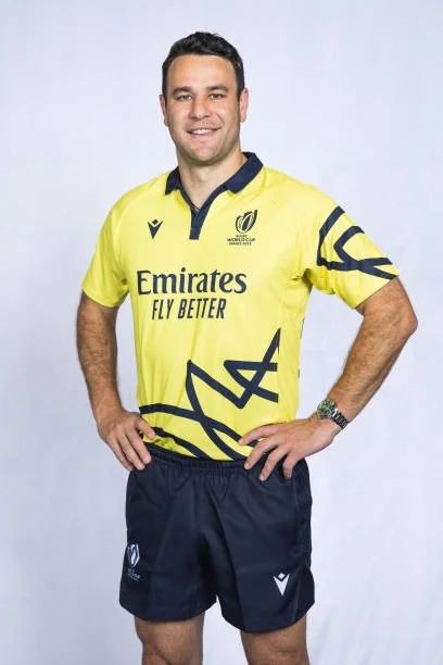 Ben O'Keeffe  in a yellow referee jersey with black accents and shorts, standing with hands on his hips, smiling before the 2023 rugby world cup.