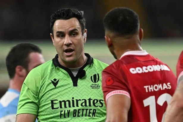 Referee Ben O'Keeffe in a green shirt talking to a player in a red jersey during a rugby match.