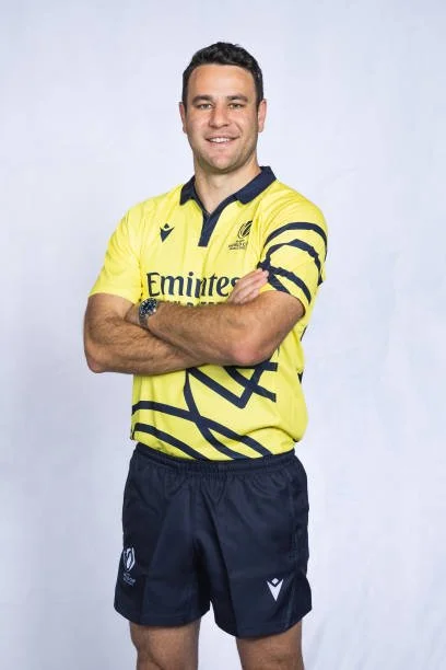 Ben O'Keeffe wearing a yellow sports referee jersey from 2023 RWC with dark blue accents and black shorts, standing with arms crossed and smiling against a light background.