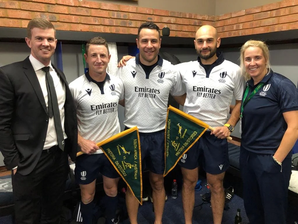 Group of five referees, three men in rugby uniforms holding flags, one man in a suit, and one woman in a team shirt, standing in a room with brick wall background. Including Ben O'Keeffe referee.