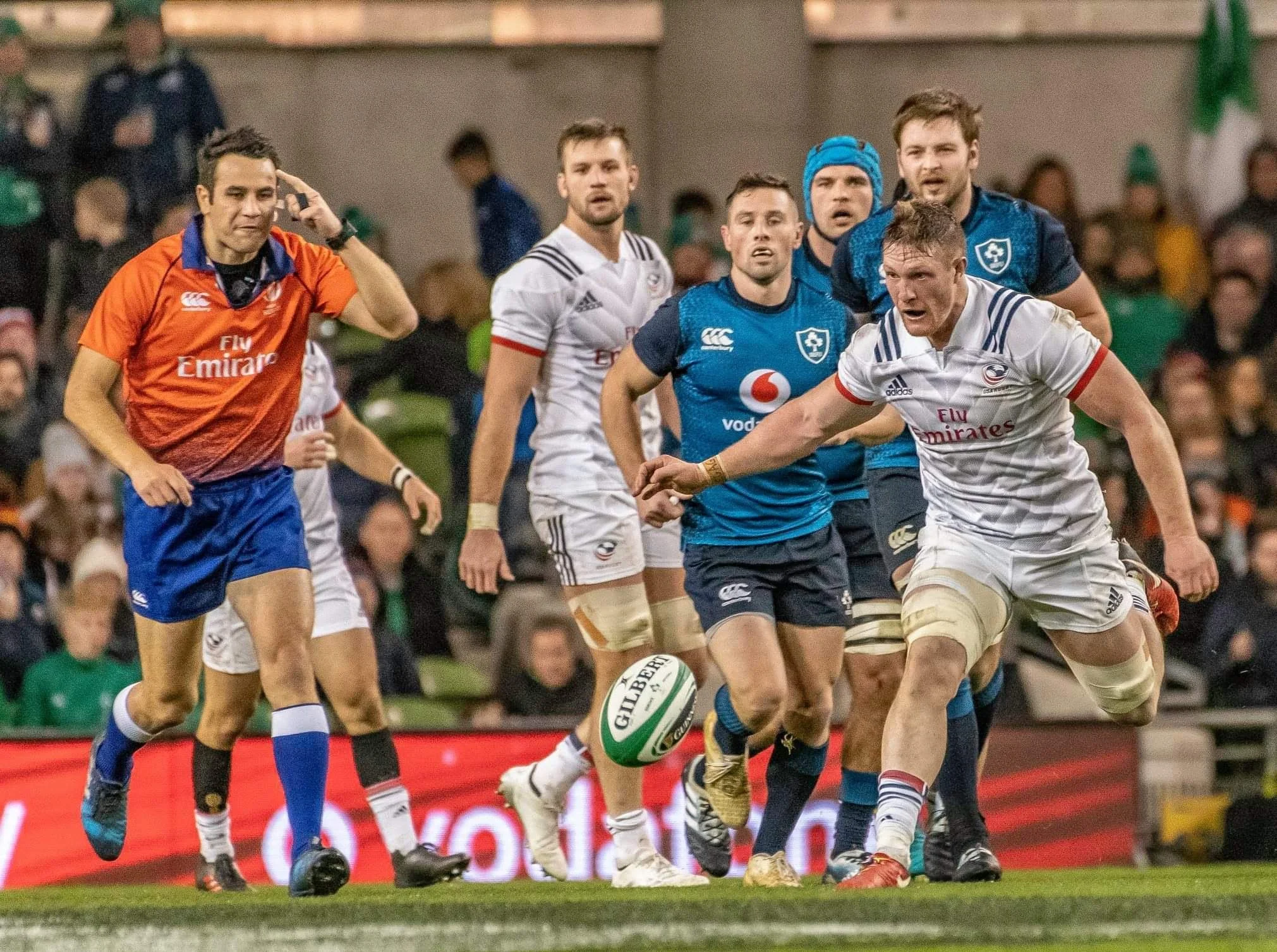 Rugby players competing for the ball on the field, with referee Ben O'Keeffe and spectators in the background.