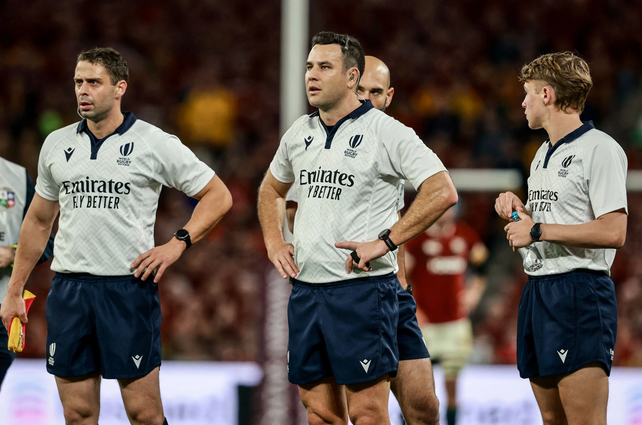 Four rugby referees including Ben O'Keeffe in white jerseys and navy shorts standing on a field with a crowd in the background during Lions series in Australia.