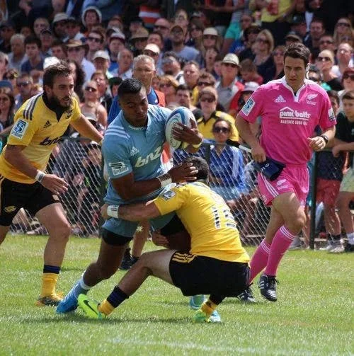 Rugby match with players in yellow and blue uniforms, one player tackling another in yellow, in front of a crowd of spectators and a referee Ben O'Keeffe .