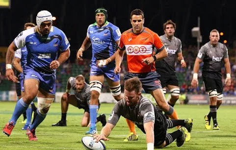 Rugby players on the field during a match, with one player diving to score a try and others chasing and referee Ben O'Keeffe.