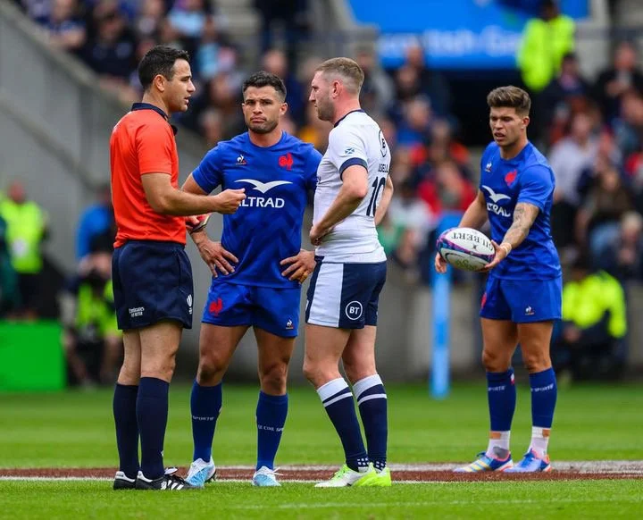 Four rugby players and referee Ben O'Keeffe on the field during a game, with a crowd watching from the stands.