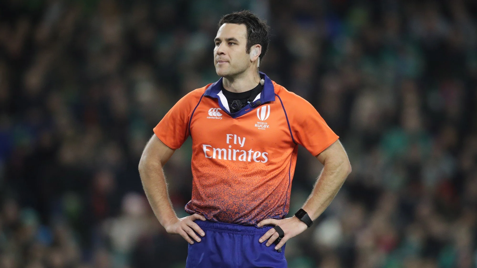 A rugby referee Ben O'Keeffe wearing an orange and blue uniform with the words 'Fly Emirates' on the front, standing on a rugby field with hands on hips and a focused expression.