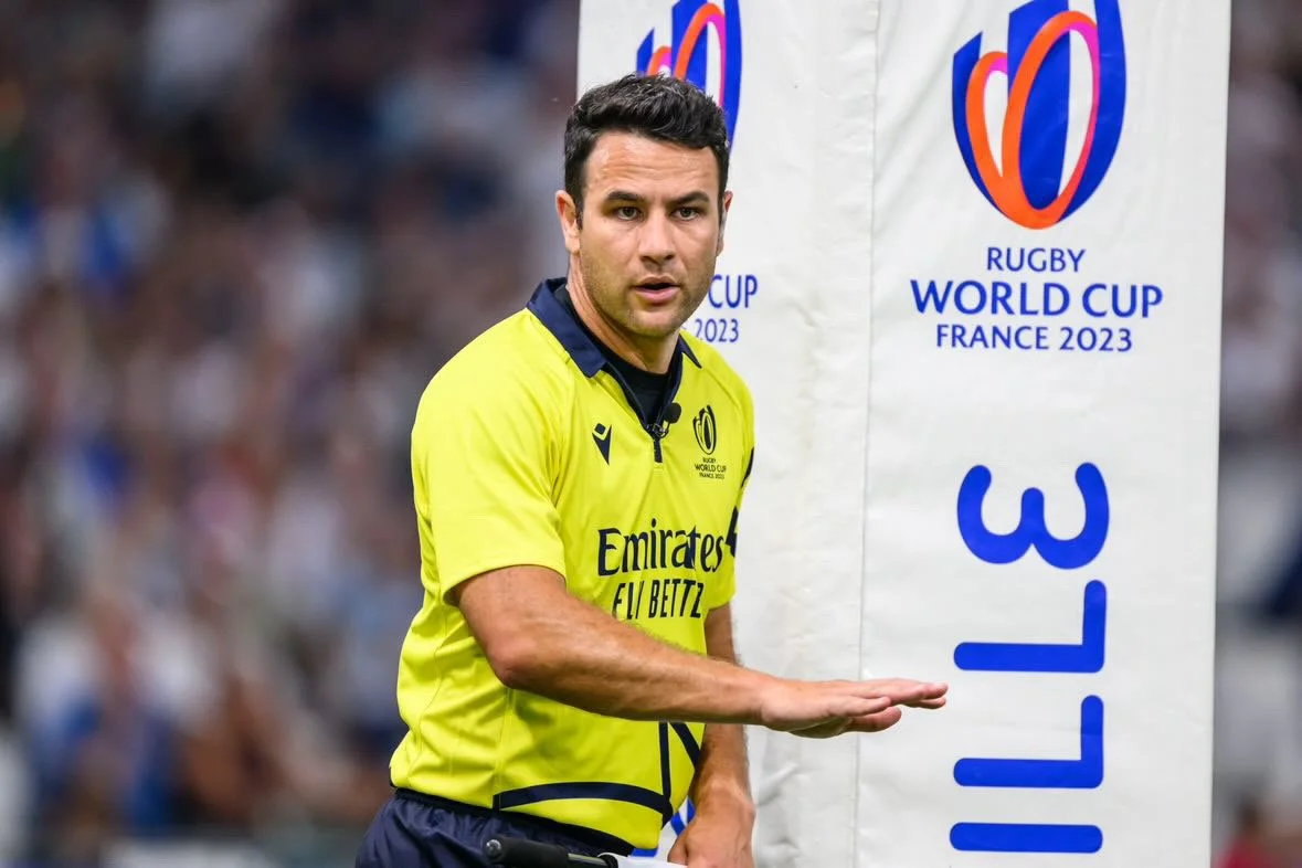 Ben O'Keeffe, a rugby referee wearing a yellow shirt with black and blue details gestures with his right hand during a game at the 2023 Rugby World Cup in France. In the background, there is a sign with the Rugby World Cup logo and text.