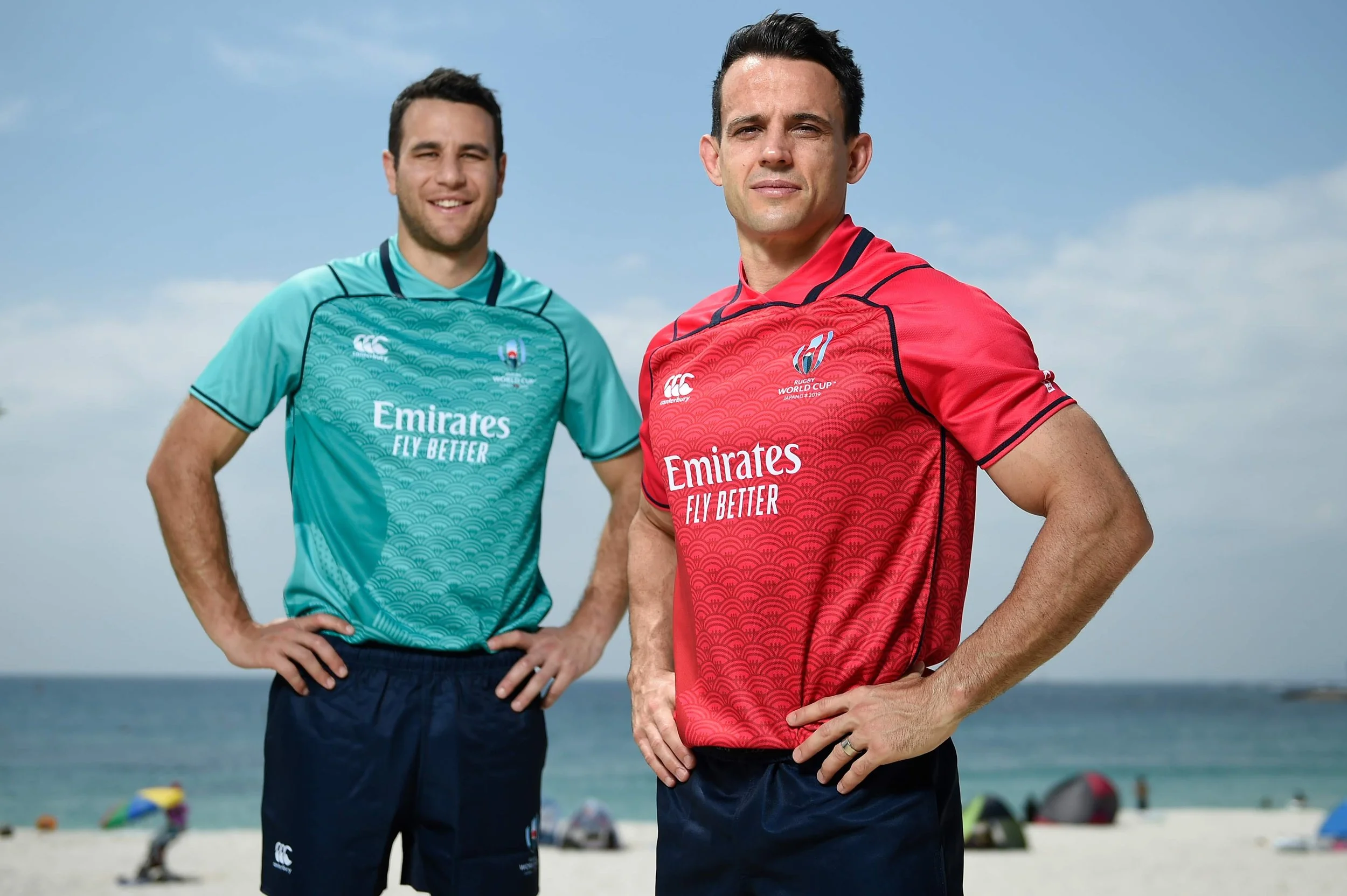 Two men stand on a beach wearing sports jerseys with the Emirates logo, one in red and the other in teal, with their hands on their hips, posing for a photo before 2019 Japan world cup including Ben O'Keeffe and Nic Berry,