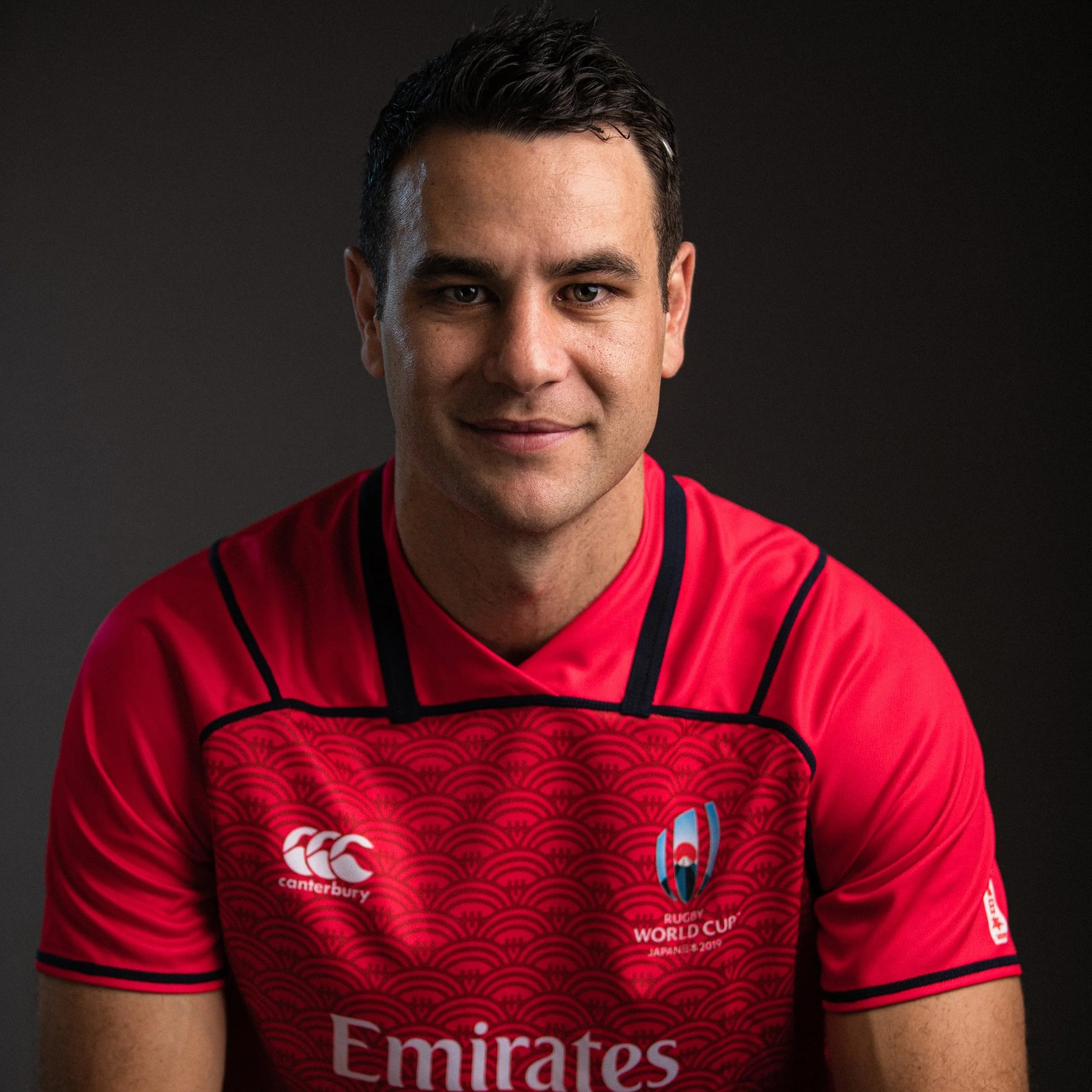Ben O'Keeffe referee is a man wearing a red rugby jersey with logos, smiling at the camera against a dark background.
