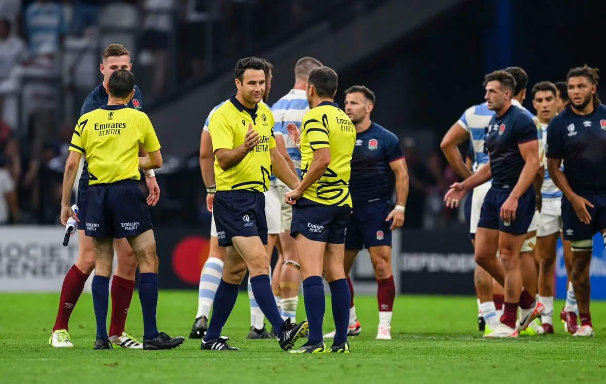 Referees on a rugby field, some wearing yellow jerseys and others in blue and white jerseys, engaged in discussion or preparing for a game including Ben O'Keeffe.