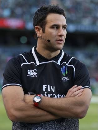 Ben O'Keeffe rugby referee wearing a black Canterbury shirt with the World Rugby logo, standing on a field with his arms crossed.