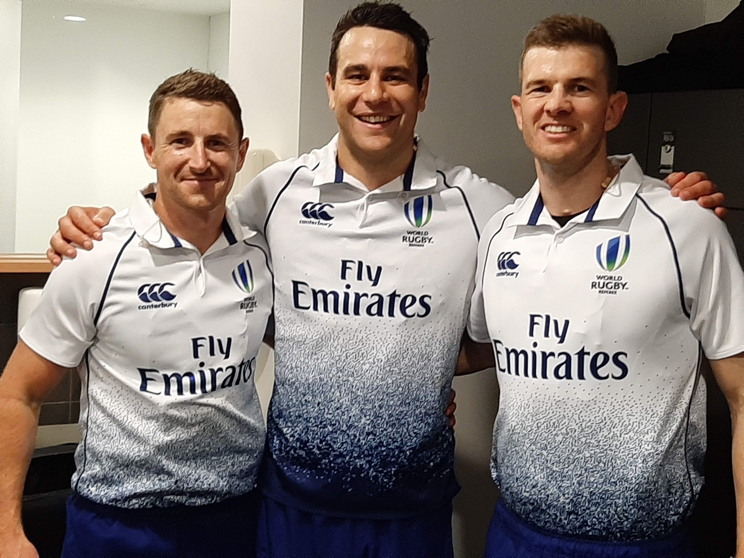 Three male referees wearing white rugby jerseys with blue and green details, posing together indoors, smiling, with arms around each other. Including Ben O'Keeffe.