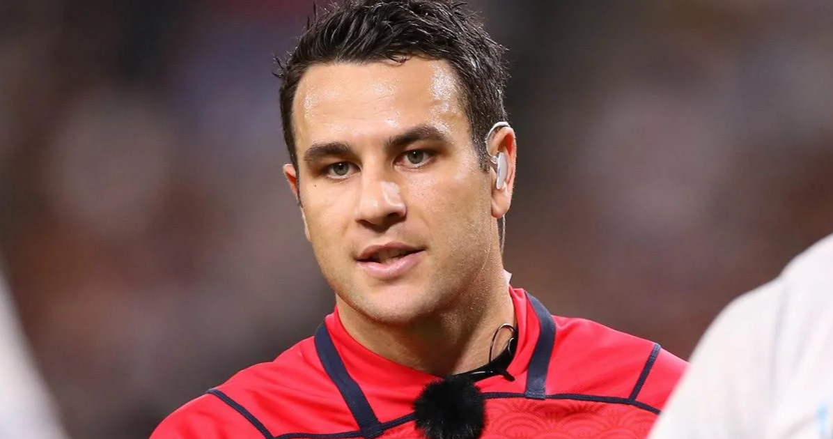 Close-up of Ben O'Keeffe with short dark hair, wearing a red sports jersey, with a microphone attached to his collar, in a sports setting.