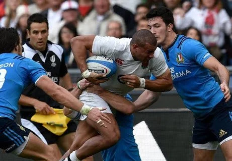 Rugby players in a match, with one player holding the ball and being tackled by opponents Ben O'Keeffe in the background.