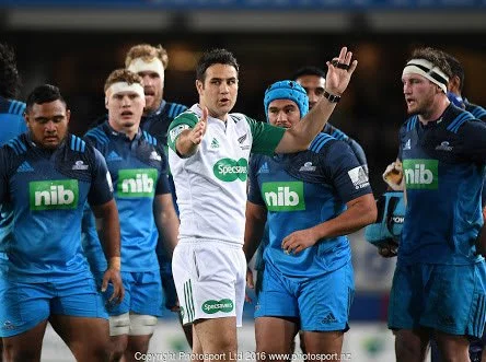 Rugby players in blue uniforms listening to referee Ben O'Keeffe in white jersey giving instructions on the field.