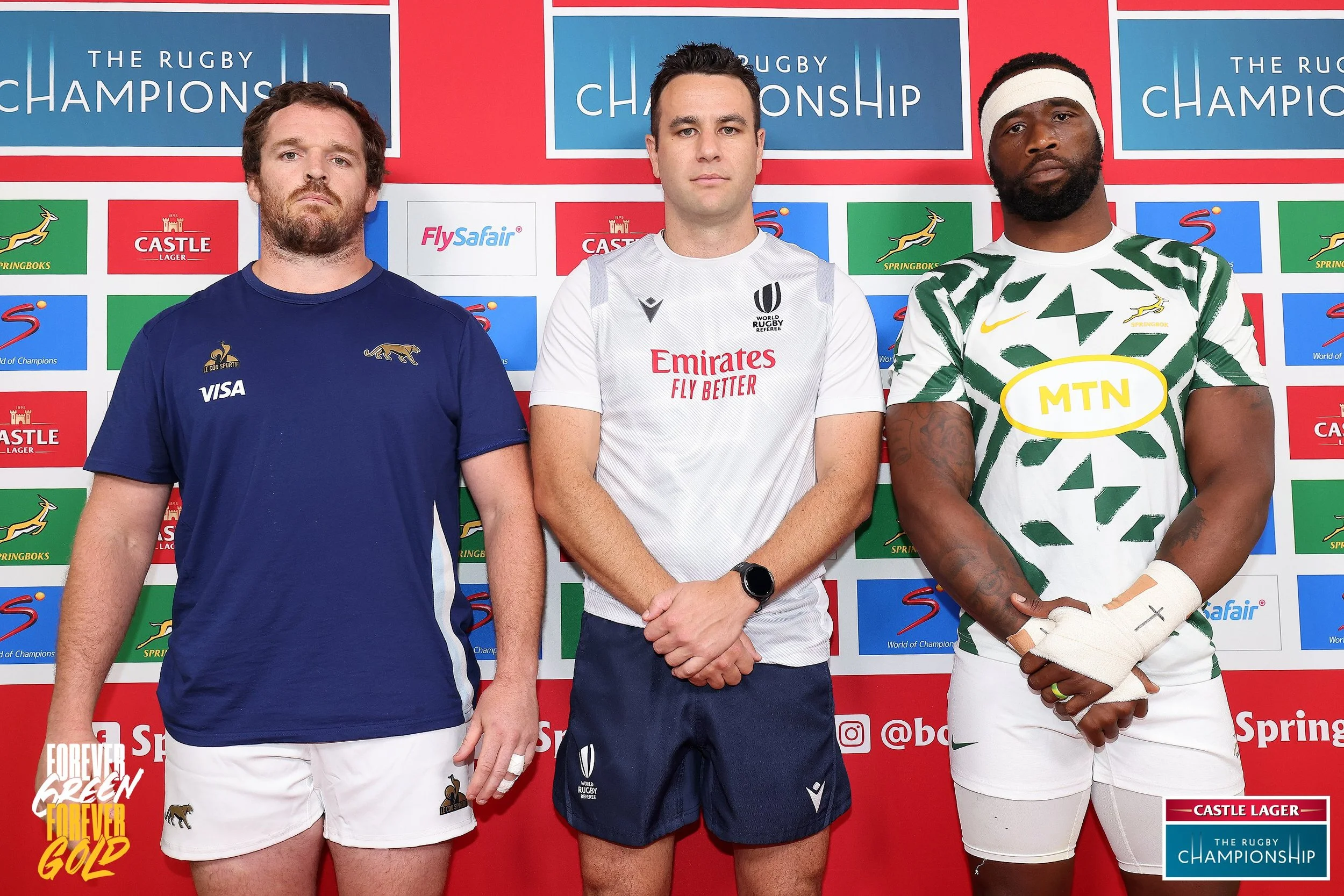 Two rugby players and referee Ben O'Keeffe standing side by side at a press event, with a backdrop featuring logos of sponsors and rugby championship branding.