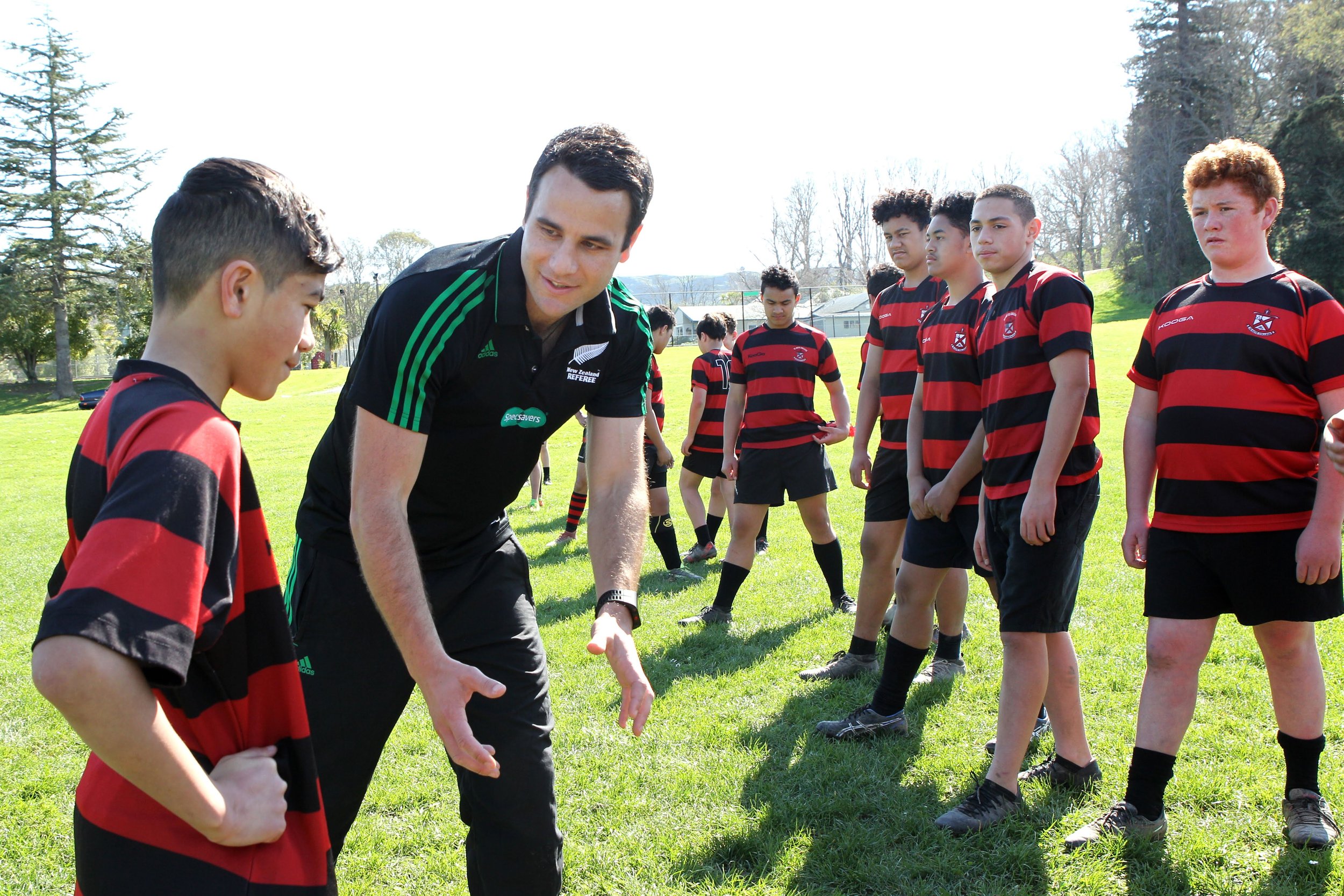 Ben O'Keeffe instructs young rugby players on a grassy field during daytime, with several players standing in line.