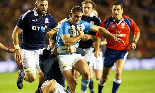 A rugby player holding a ball runs forward with multiple players chasing him on a field during a game with referee Ben O'Keeffe.
