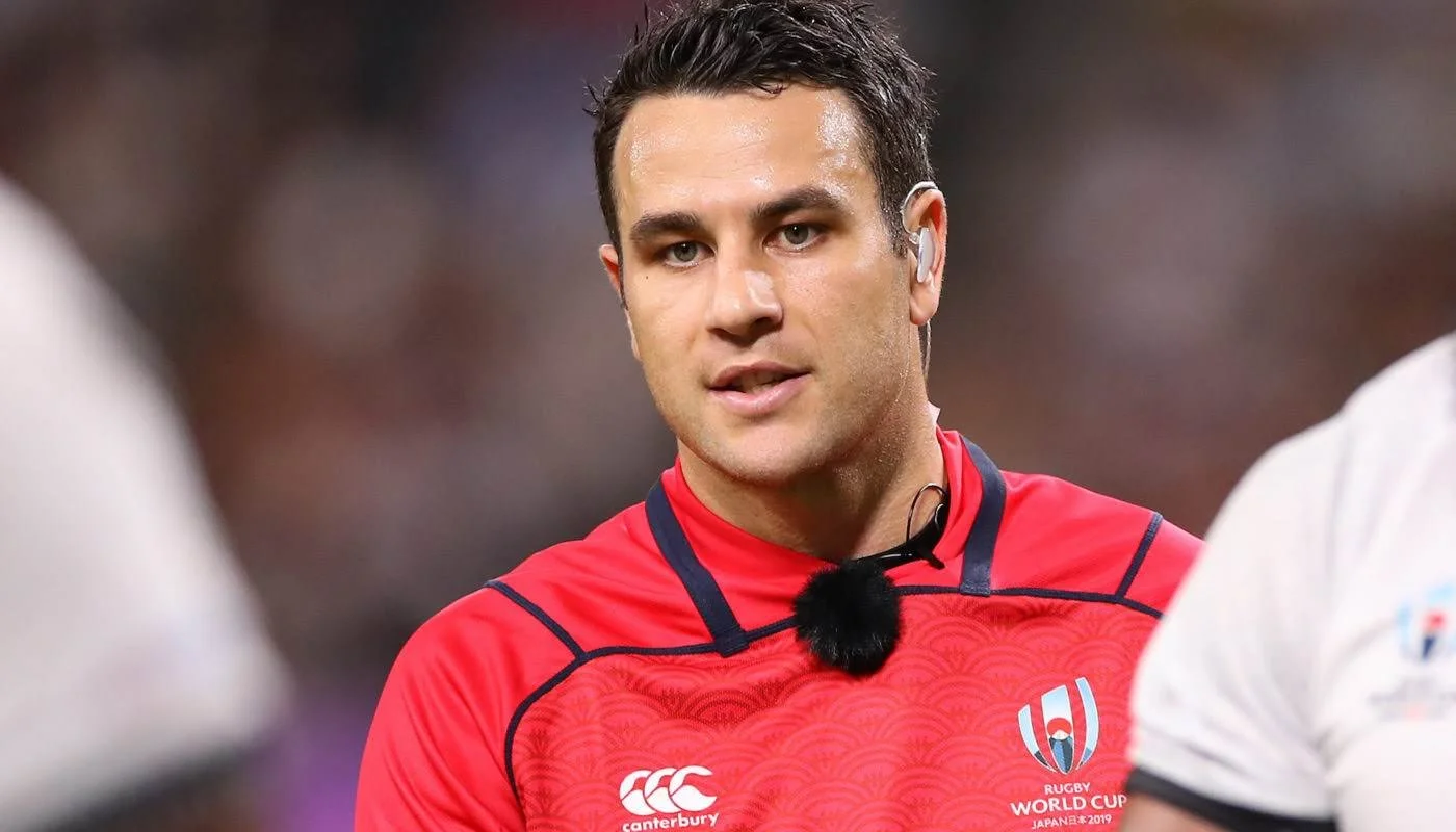 Ben O'Keeffe with dark hair wearing a red rugby jersey with the rugby world cup logo and Canterbury logo, and a black lapel mic attached to his shirt.